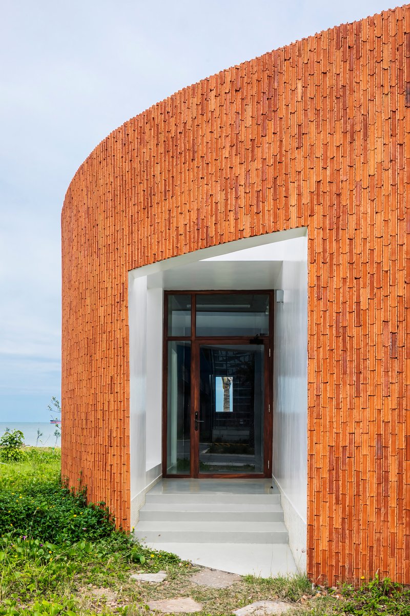 Recessed entrance with white concrete steps through curved terracotta brick wall facing coastal vegetation