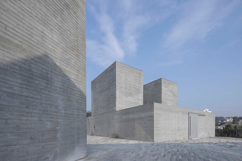 Exterior courtyard framed by board-formed concrete volumes with a stone-paved plaza beneath clear skies