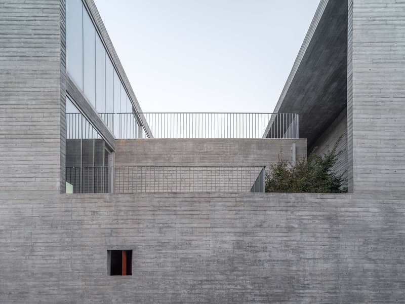 Layered grey brick walls framing an elevated courtyard with metal railing under an overcast sky