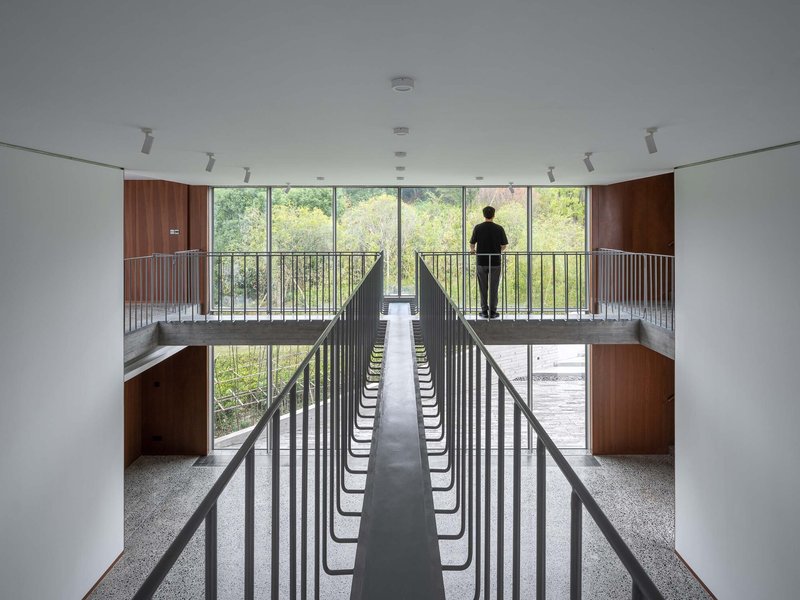 Double-height interior with vertical black railings and a figure silhouetted at the glazed balcony overlooking trees