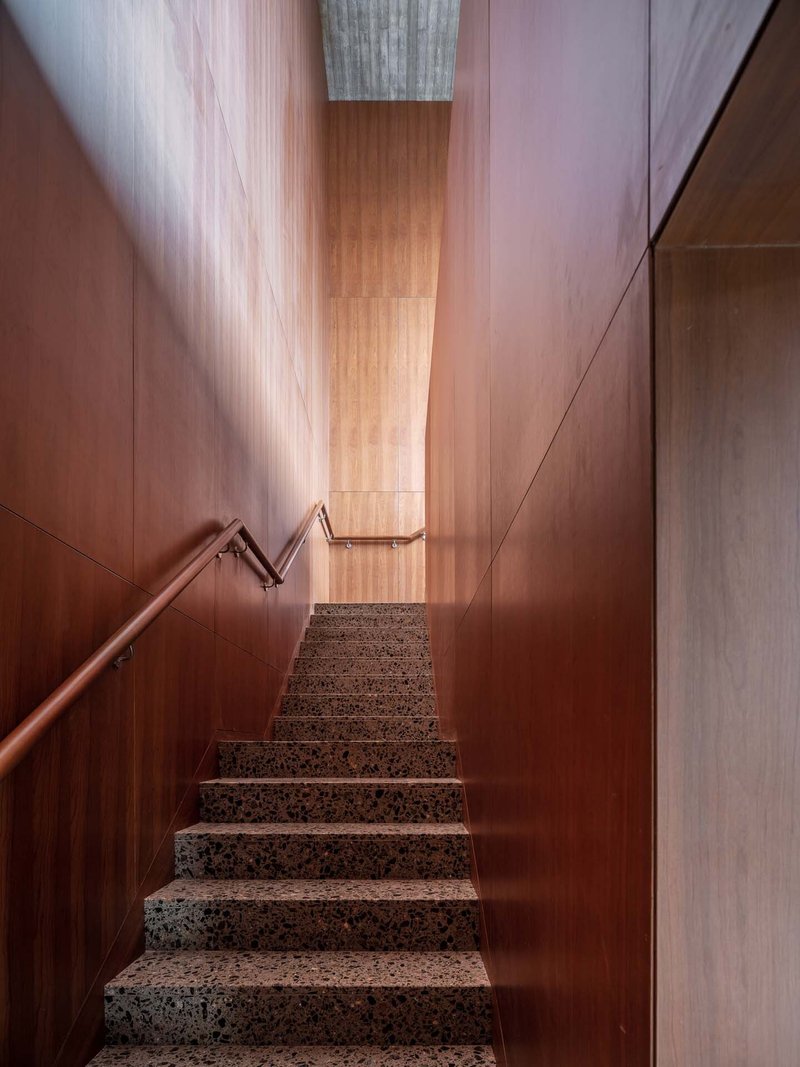 Narrow staircase with terrazzo treads and wood-paneled walls rising toward a skylight above