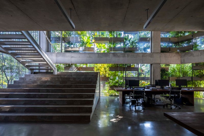 Concrete staircase rising beside open workspace with desks below planted balcony edges visible through glazing