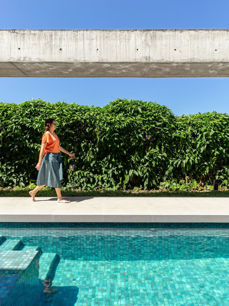 Pool deck with concrete beam overhead, dense hedge boundary, and person in orange shirt walking past