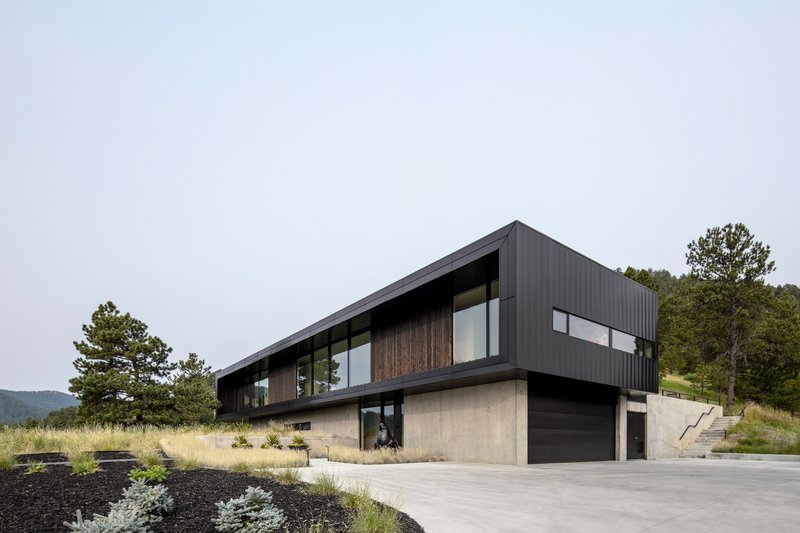 Two-story volume with black corrugated metal upper level and concrete lower level beside a concrete driveway
