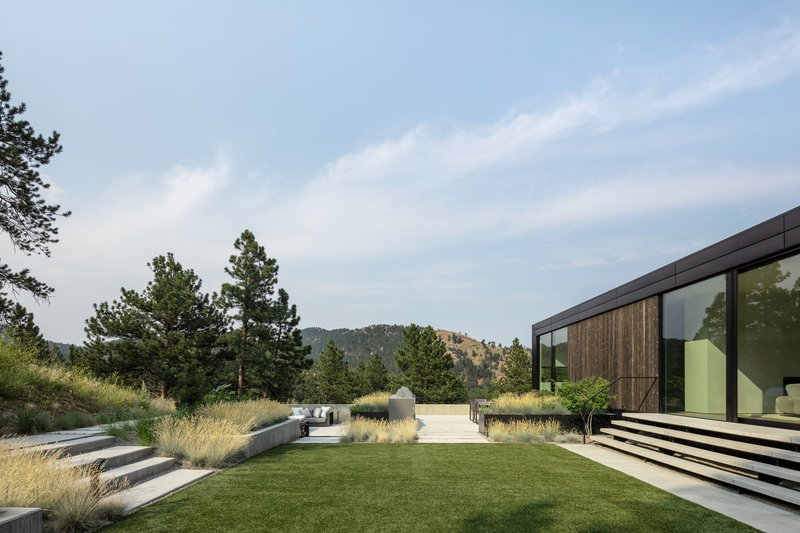 Terraced lawn and gravel pathway extend through native grasses toward forested hills beyond the glass facade