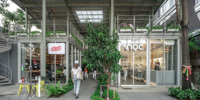 Ground-level passage with glass storefronts and mesh railings as a visitor walks through planted greenery