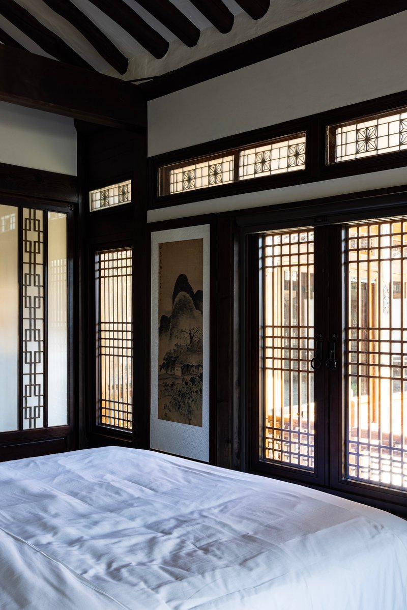 Corner of a bedroom showing latticed screens, a vertical scroll painting, and filtered daylight