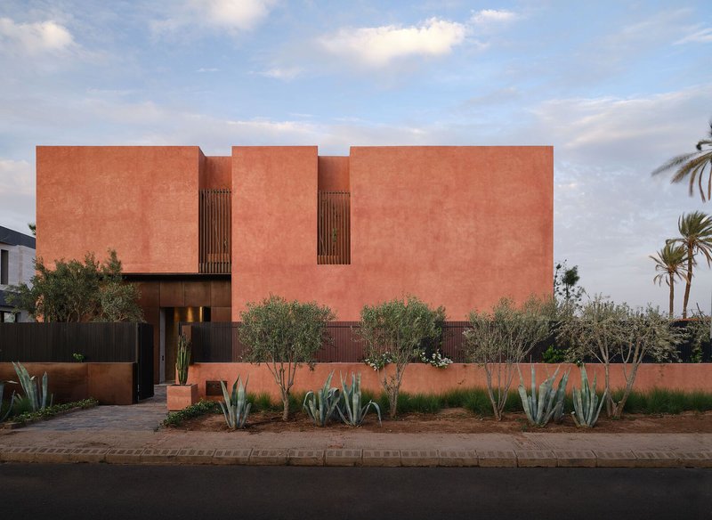 Street view of terracotta-rendered facade with vertical timber louvers and planted agave bed at dusk