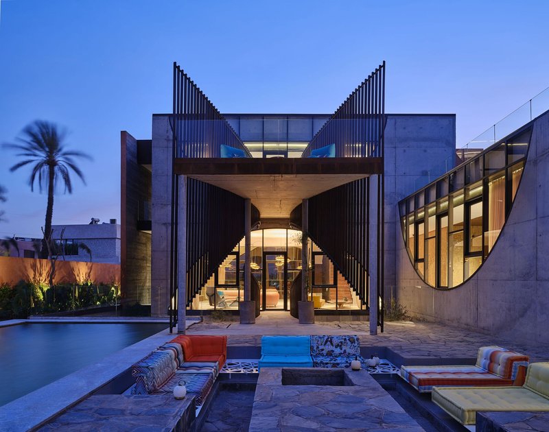 Rear courtyard with concrete volumes, arched opening, and vertical louvered towers illuminated at twilight