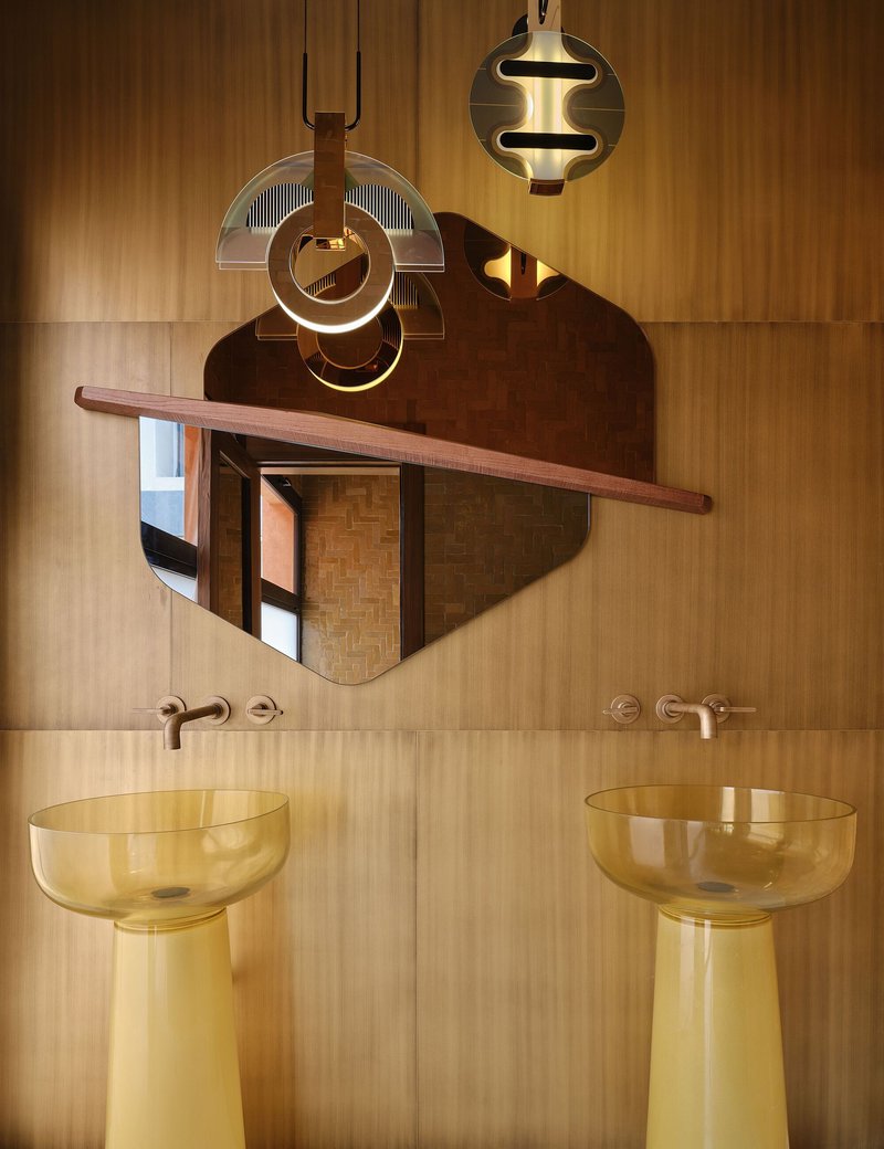 Bathroom vanity with twin yellow glass pedestal sinks beneath sculptural pendant lights and a curved timber shelf mirror
