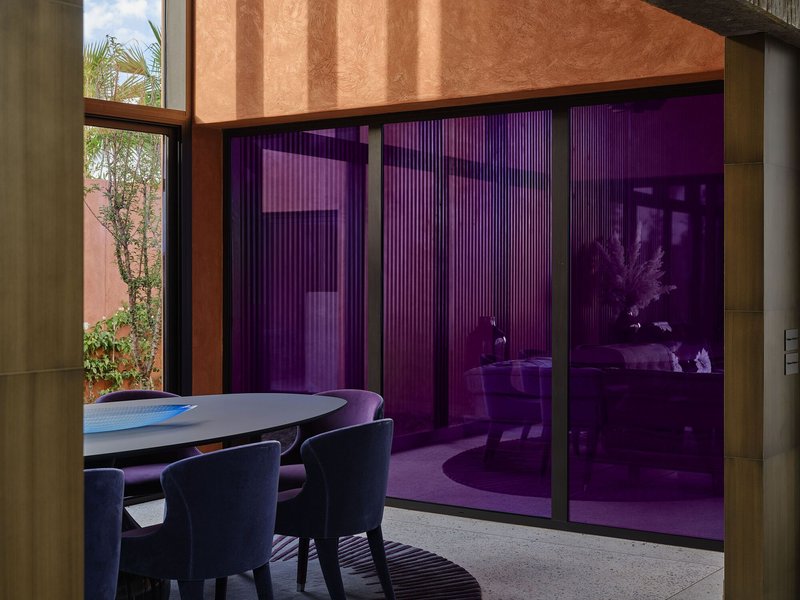 Dining space with purple-tinted glass wall separating interior from courtyard with planted bed in afternoon light