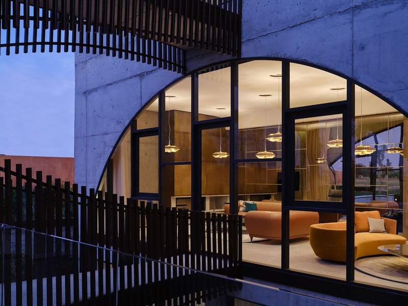 Arched glazing set within concrete walls framing an illuminated interior lounge at dusk
