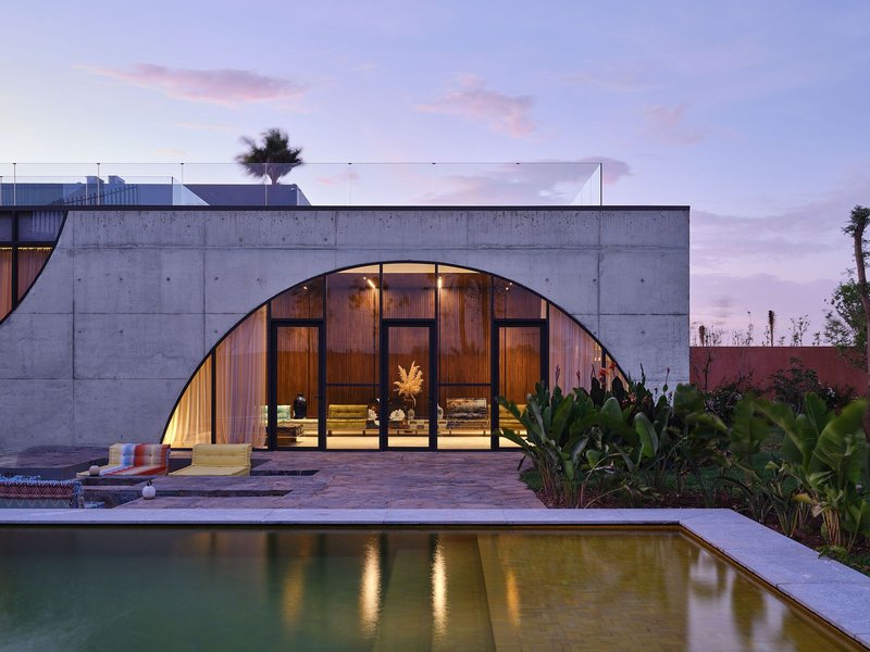 Concrete facade with arched glazed opening facing a pool and tropical plantings at twilight