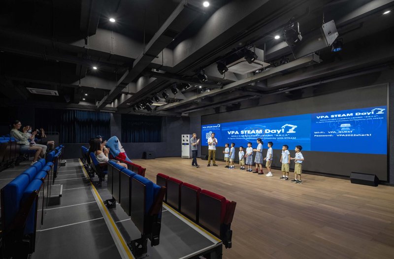 Auditorium with tiered seating and exposed black ceiling facing a blue projection screen during a presentation