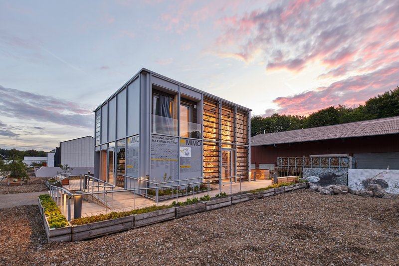 Two-storey corrugated metal structure with timber screen facade and planted beds under a pink sunset sky