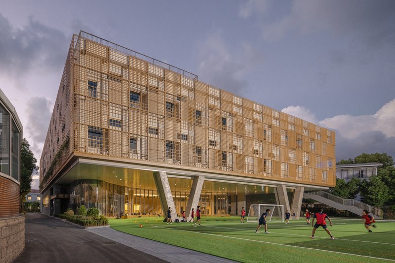 Perforated metal facade above glazed ground level with students on synthetic turf at dusk