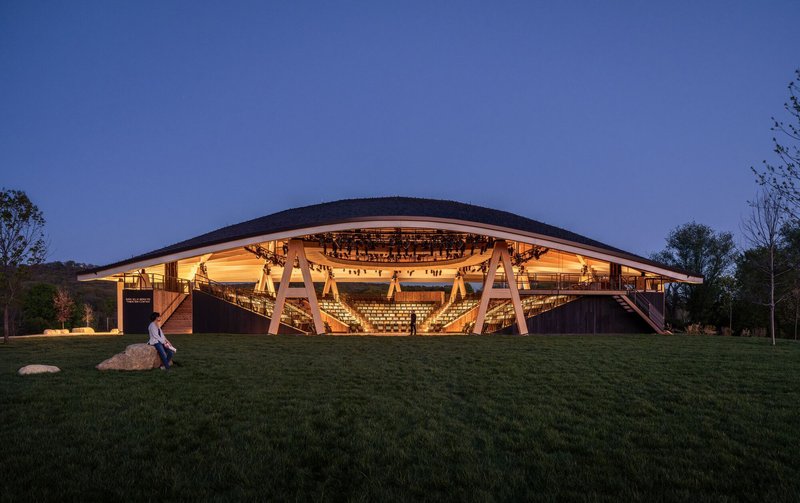 Illuminated facade with exposed timber trusses and amphitheater seating visible through the glazed perimeter at dusk