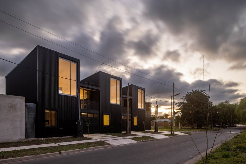 Facade of black corrugated townhouses with glowing interiors visible through tall windows at twilight