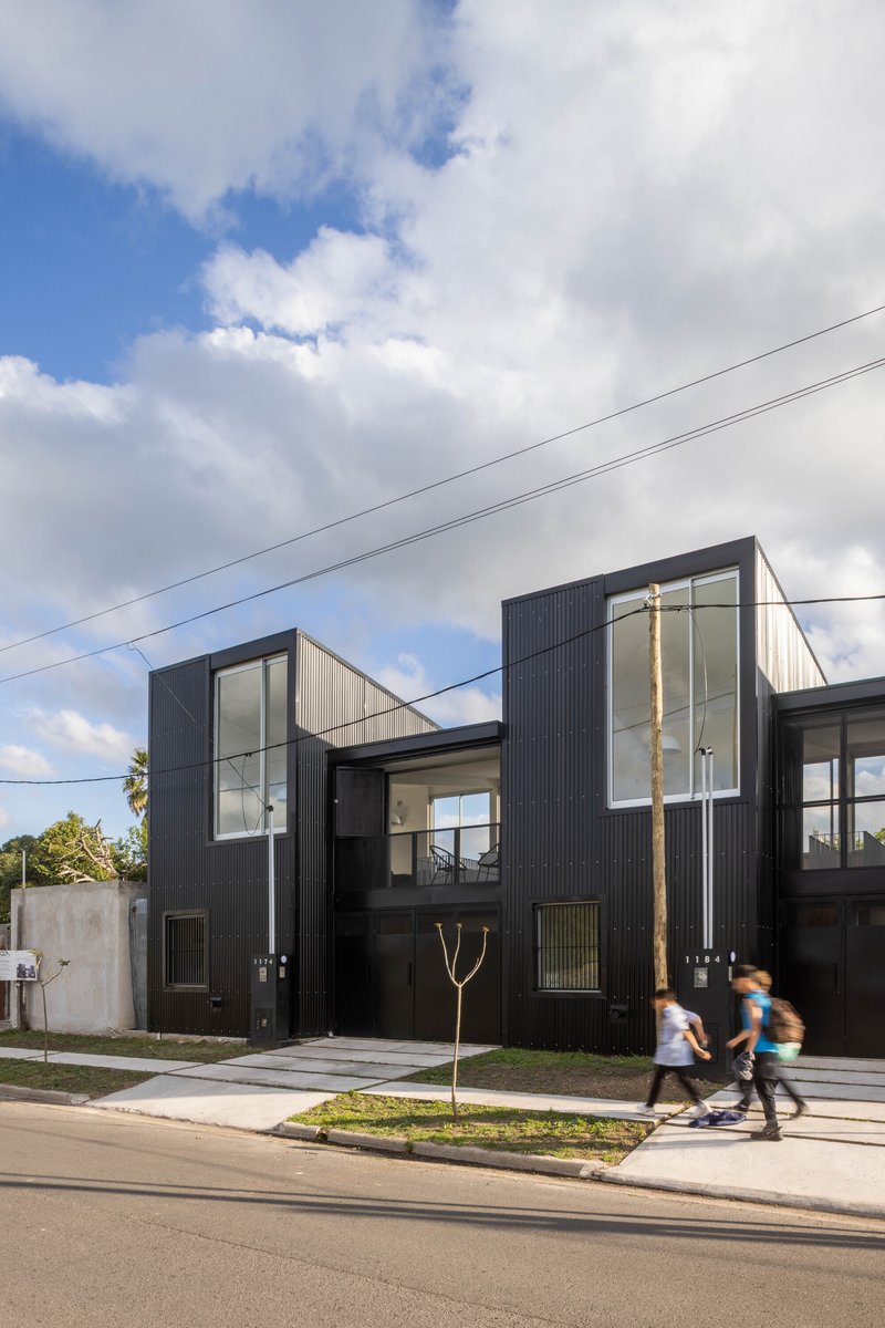 Street view of the black corrugated metal facade with glazed volumes and two people cycling past on the sidewalk