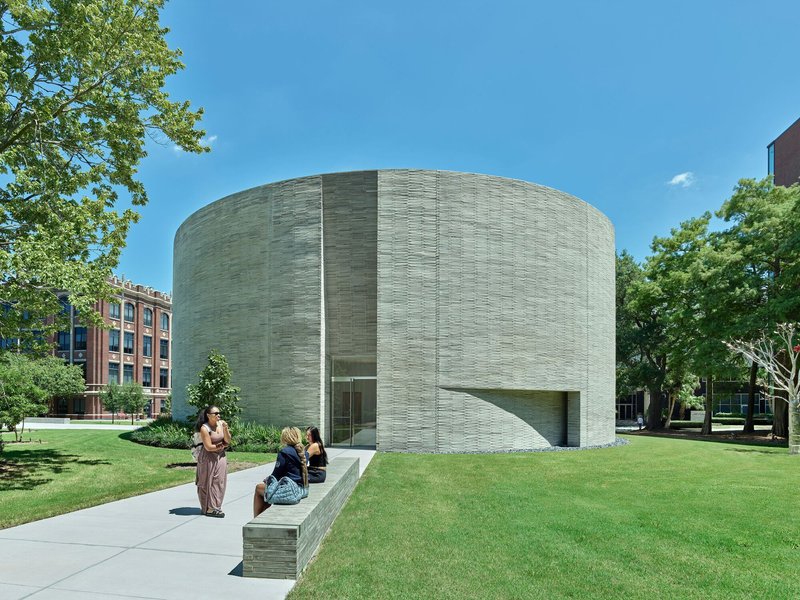 Cylindrical building with horizontal striped masonry facade set in a lawn with visitors on a bench