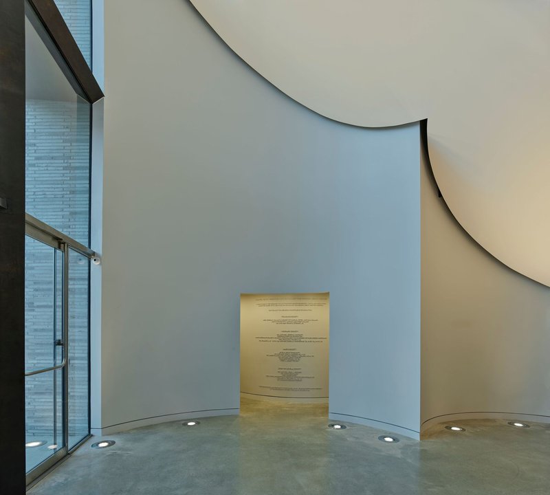 Curved interior wall with narrow illuminated doorway and floor-recessed uplights beside glazed facade
