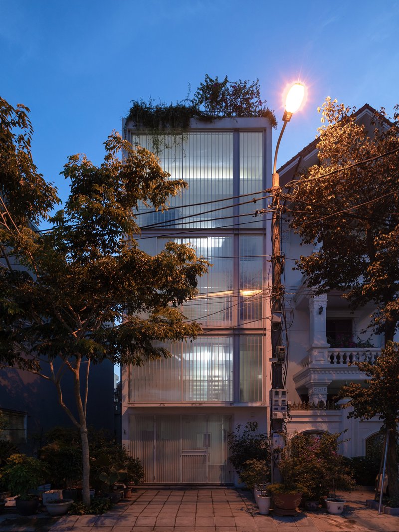 Narrow tower facade with corrugated polycarbonate panels glowing at dusk behind overhead power lines and trees