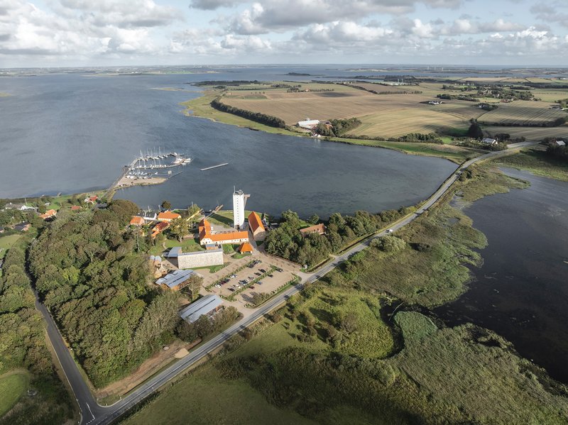 Aerial view of a peninsula with red-roofed buildings and white tower nestled among trees at the water's edge