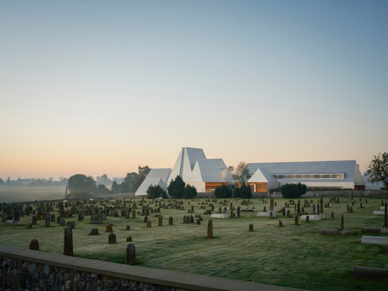 White faceted roof volumes rising above a cemetery at dawn with mist covering distant fields
