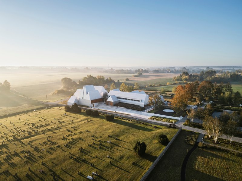 Aerial view of angular white roofs and linear wings bordered by a cemetery and morning fog