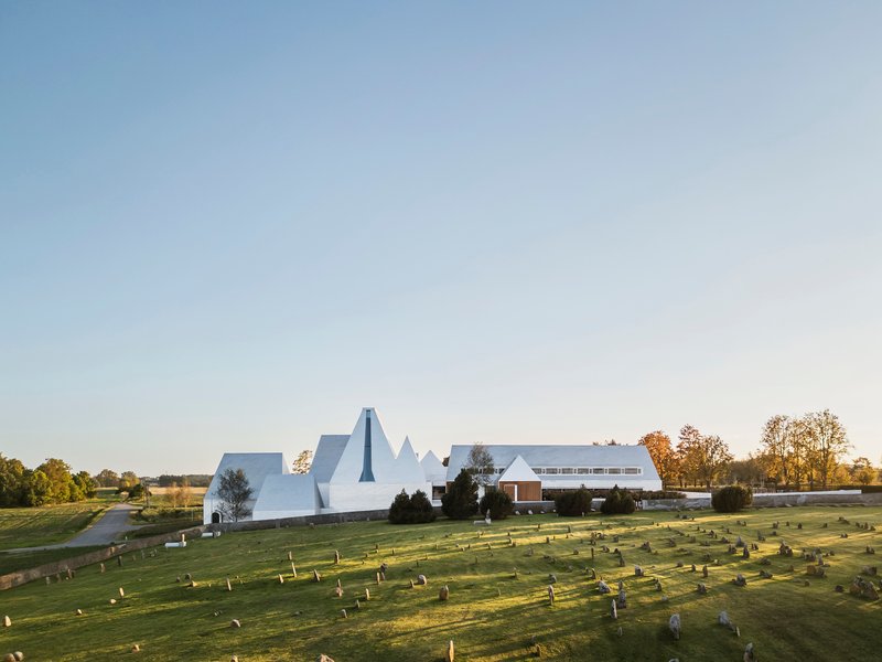 Clustered peaked white roofs and horizontal wing overlooking a grassy burial ground in afternoon light