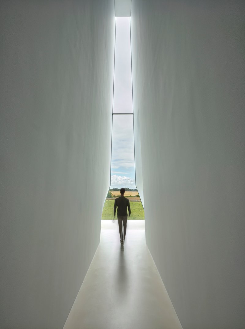 Tall vertical window slot framing a silhouetted figure walking toward countryside beyond white walls