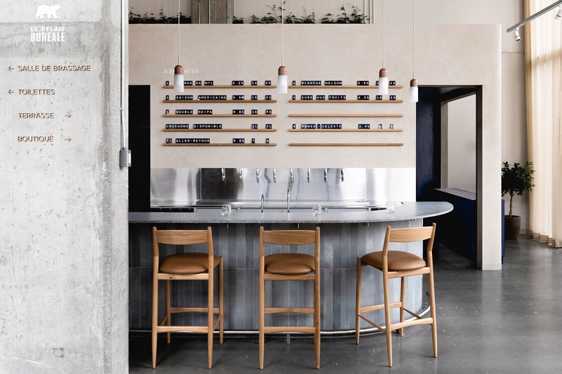 Curved bar with fluted tile base, oak stools, and wall-mounted shelving beside concrete directional signage panel