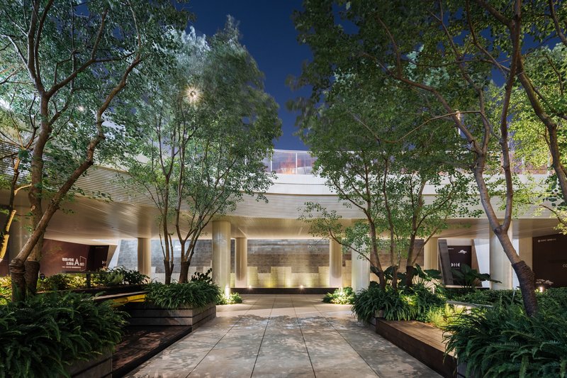 Evening view of illuminated entry pavilion with stone walls and uplighting beneath established trees at dusk