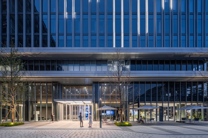 Ground level plaza with glass entrance pavilion, paired figures walking, and young trees at dusk