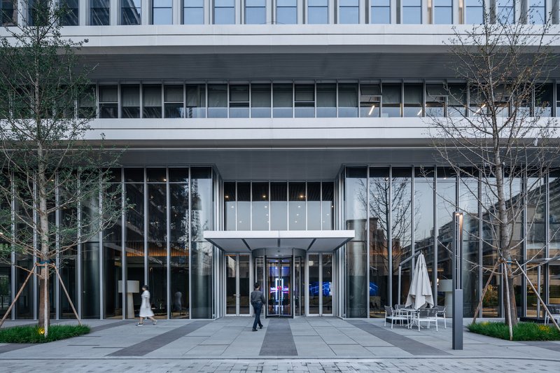 Entrance canopy with glass walls flanked by young trees and a person walking in early evening