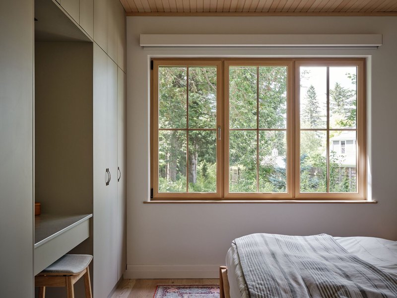 Bedroom with timber-framed casement windows overlooking summer foliage and neighboring houses