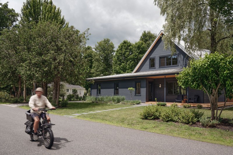 Street view of dark timber-clad gabled house with cyclist passing under cloudy sky