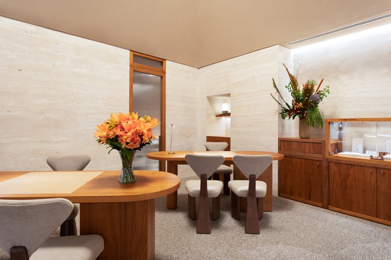 Interior retail space with rammed earth walls, walnut cabinetry, and circular tables with vases of flowers
