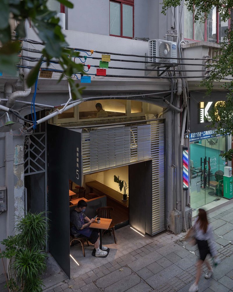 Two-story shopfront with horizontal louvers and outdoor seating as pedestrians pass in early evening