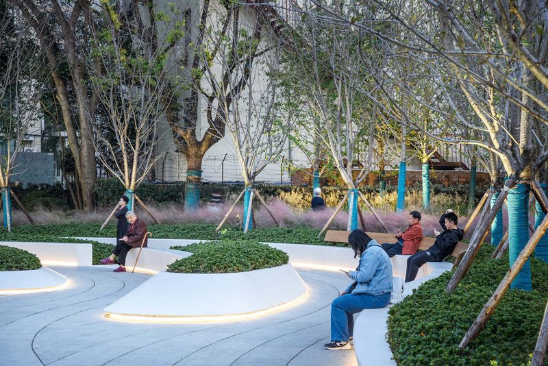 Curved white concrete benches with integrated lighting among planted beds and bare winter trees at dusk