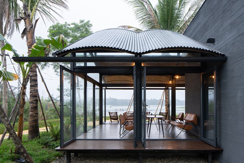 Glazed pavilion with corrugated metal roof and black steel frame nestled among palm trees overlooking the water