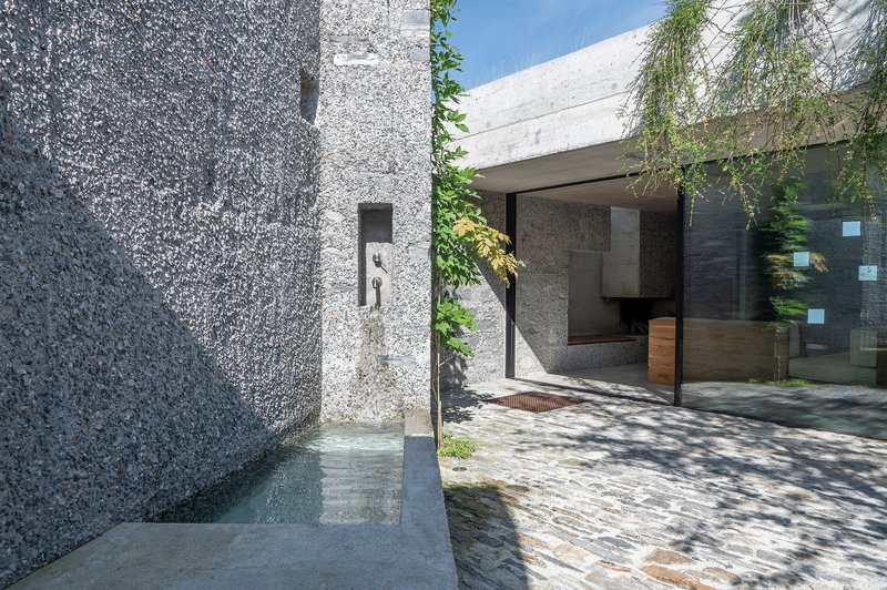 Narrow entry corridor between textured concrete walls leading to glazed interior space with trailing vines overhead