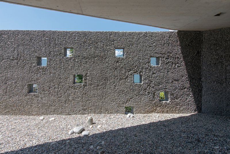 Exterior gravel courtyard wall with scattered square window openings beneath concrete overhang