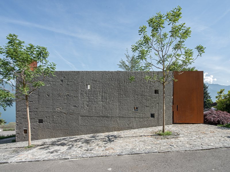 Street view of textured concrete facade with scattered windows and a weathered steel volume under a clear sky