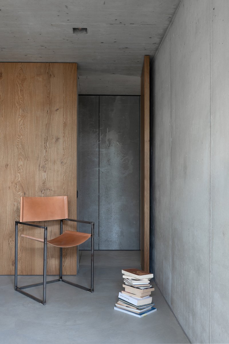 Interior corner with leather chair, stacked books on the floor, and timber cabinetry against concrete walls