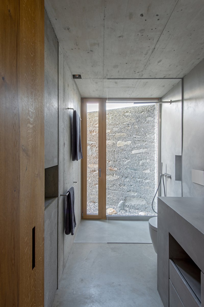 Bathroom with concrete walls, timber cabinetry, and a narrow window facing the textured courtyard wall