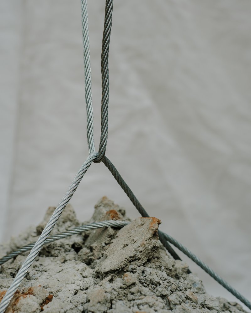 Steel cables securing a rough concrete block suspended against a white fabric backdrop