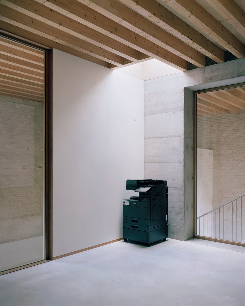 Corner nook with exposed timber joists overhead and printer stationed against white partition near glazed opening