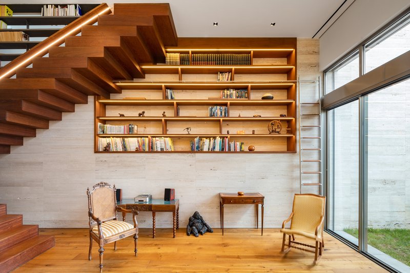 Floating timber staircase beside built-in bookshelves with integrated lighting and tall glazing to a courtyard