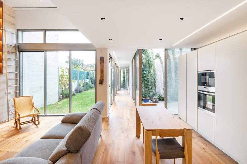 Long interior corridor connecting living and kitchen zones with planted courtyards visible through floor-to-ceiling glass